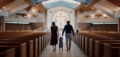 A woman and man hold hands with a toddler in between them while walking down the center aisle of a nearly empty church filled with natural light.