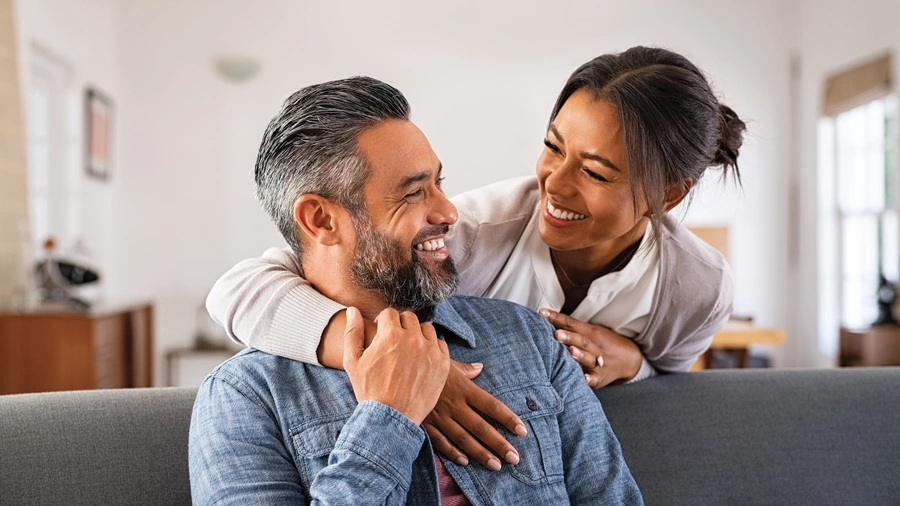 Smiling woman hugging her husband on the couch from behind in the living room