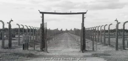 A black and white video still of a gate to a concentration camp.