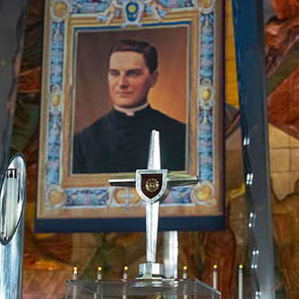 A portrait of Blessed Michael McGivney is surrounded by a relic and several fancy candles in silver candle holders.
