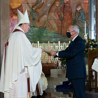Cardinal Joseph Tobin dressed in a white priest gown and hat stands in front of Carl Anderson who is dressed in a black suit.  In the background, 4 other priests can be seen in white priest garb, one standing and three seated.  Artwork can also be seen in the background.