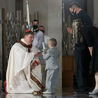 Young child wearing a light grey suit approaches Cardinal Tobin with a relic in hand to present to him. Onlookers can be seen in the background.