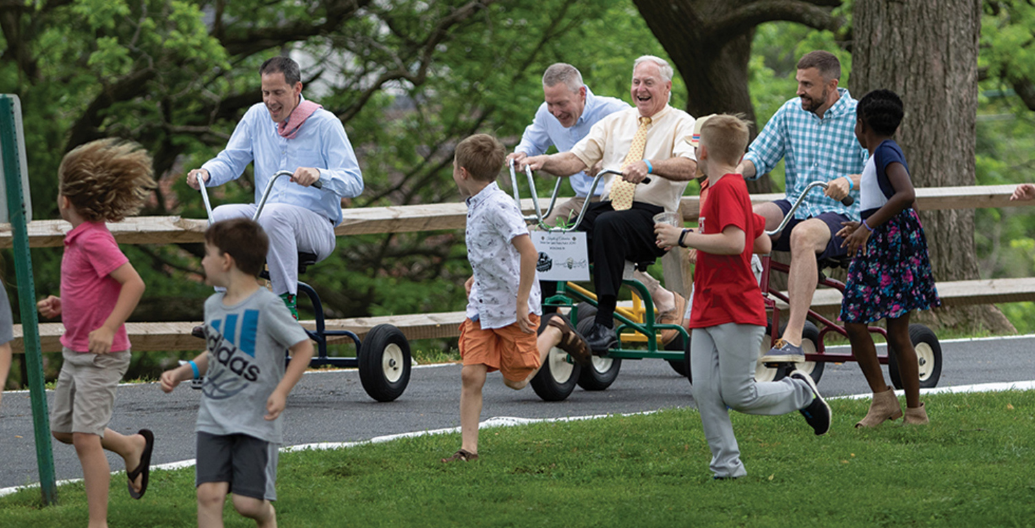 Adults riding on tricycles and children are running with them.
