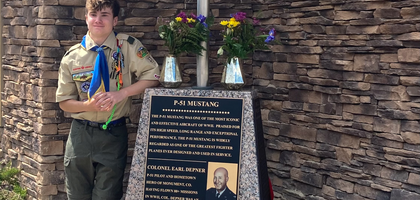 Colin Saber, a Life Scout with Troop 17, dressed in his Life Scout attire, stands outdoors posed up against a P-51 Mustang sculpture.