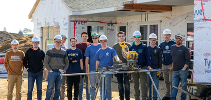 Young adults dressed in casual clothing and hard construction hats stand outdoors with tools outside of a Habitat for Humanity build.