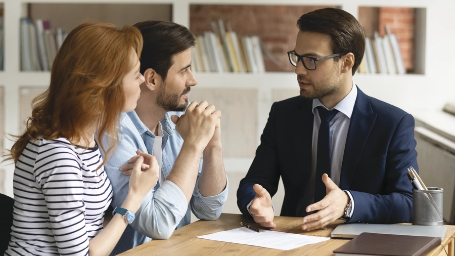 Insurance agent in a suit and tie discussing life insurance options with a married couple at desk.