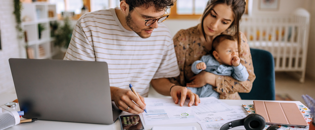 A husband and wife with a young infant sitting at a desk overviewing financial documents.