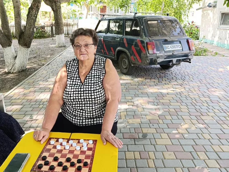 a women outdoors playing checkers sitting at a table with a black automobile visible in the background