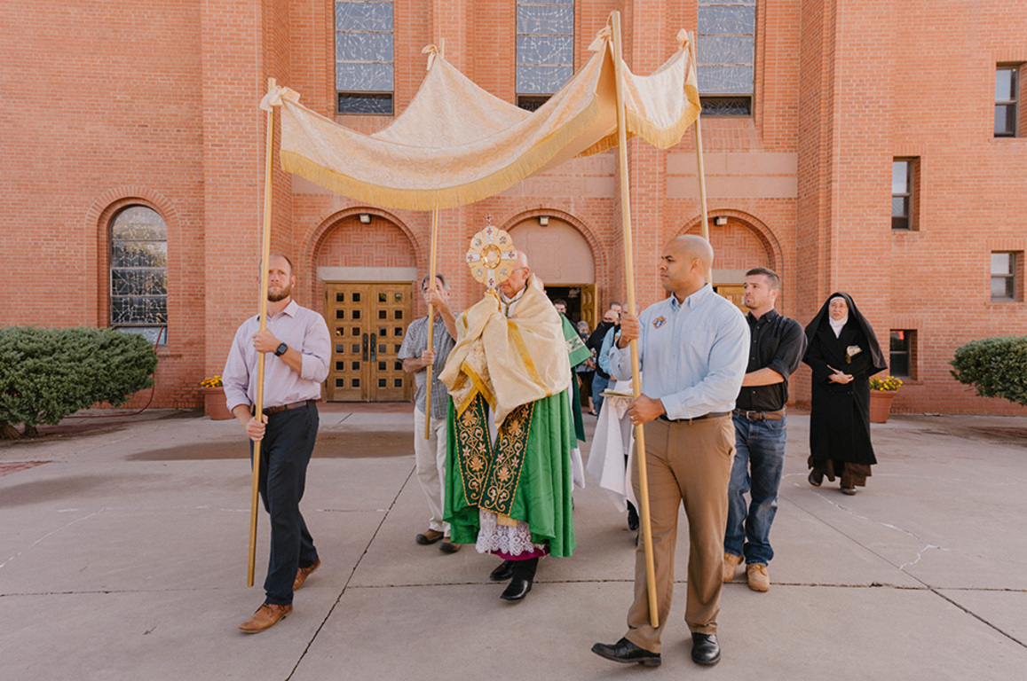 Catholic priest processes with the Eucharist in a monstrance while four men hold a processional canopy over him.