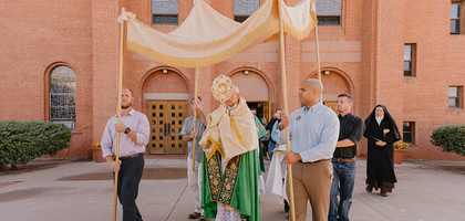 Catholic priest processes with the Eucharist in a monstrance while four men hold a processional canopy over him.