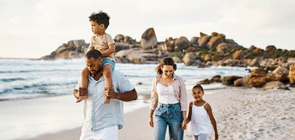 A family walking on the beach with a child on the dad's shoulders and a woman holding her daughter's hand.