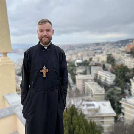 Priest standing outdoors in a black robe with a cross necklace