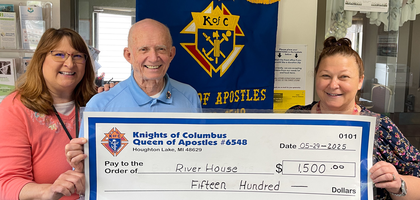 Grand Knight Clyde Straub of Queen of Apostles Council 6548 dressed in a blue Knights of Columbus polo shirt smiles alongside two women dressed in casual attire holding a large check in the amount of $1,500.