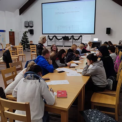 A group writes at a large, long table with a projector screen in the background.