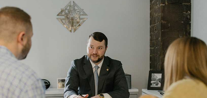 A family sitting down talking with an investment consultant.  Investment consultant is wearing a suit and sitting at a desk with paperwork in front of him and a pen in hand.  Family is a man, woman and young baby.