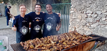 Three Knights wearing navy blue Knights of Columbus aprons and holding grilling utensils stand together behind a grill filled with cooked chicken and sausages.