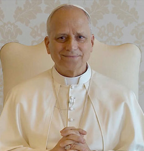 Pope Leo XIV poses for a photo seated in a tan chair and folding his hands.