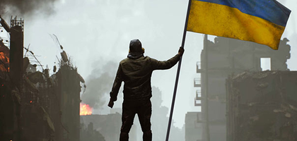 Soldier holding a Ukrainian flag on top of a pile of rubble and burning town in the background.