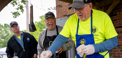 Men dressed in aprons and Knights of Columbus attire smile as they prepare food on the grill.