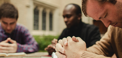 Three men bowing in prayer while reading their Bibles. One man is in focus while the other two are in the background, blurred.