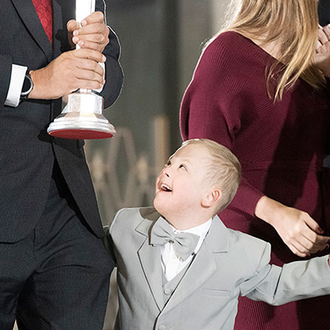 Young child wearing a light grey suit smiles as he walks alongside his father who is wearing a black suit.  His father is holding a relic in hand.