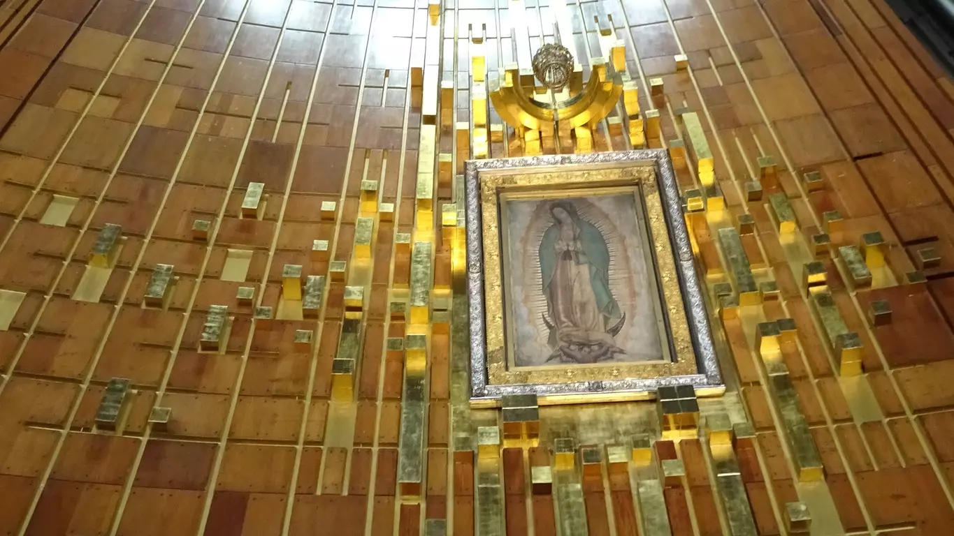 An Our Lady of Guadalupe mosaic embedded high up within a churches wall, beneath a cross, also embedded into the wall, with light shining down on both from above.