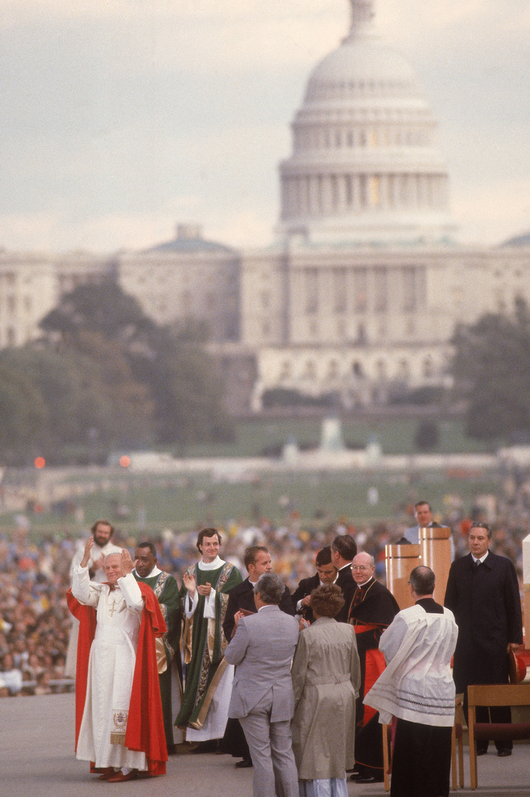 Pope John Paul II stands with his arm raised and beside a crowd of people and the US capitol building in the distance. 