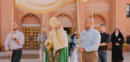 A priest holds a golden monstrance; men carry a canopy to cover the procession.