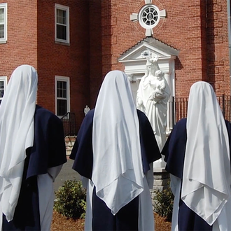 Three sisters stand outside with their backs to the camera. They are in front of a statue of the Virgin Mary holding baby Jesus.