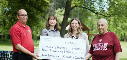 Two women standing in between two men, both wearing red shirts and slacks and holding up a large paper check in the amount of $9,000 endorsed to Mary's Mantle.