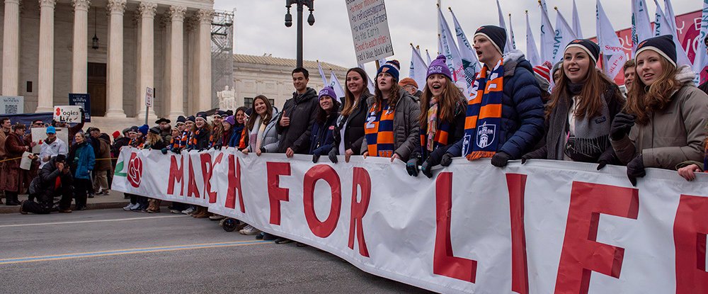 College students wearing winter coats and hats hold a white banner with the words March for Life printed on it in the street in protest.