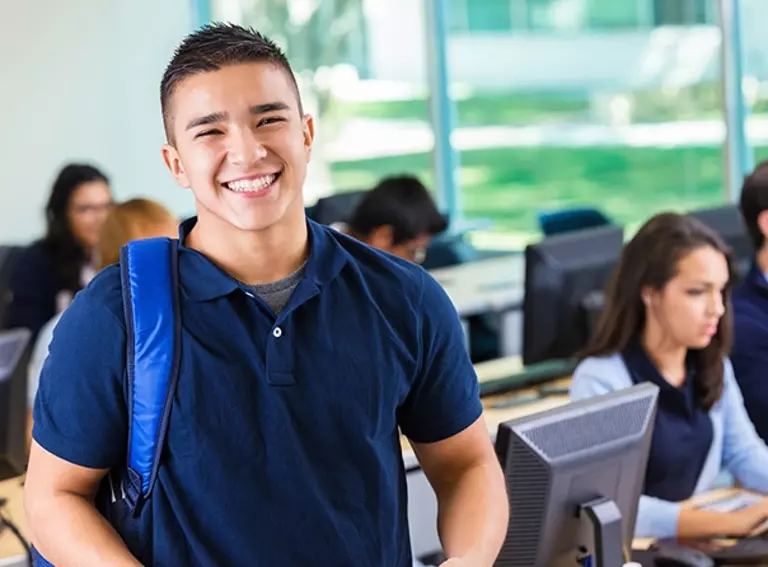 A young man smiles while standing in front of multiple computers in a classroom.