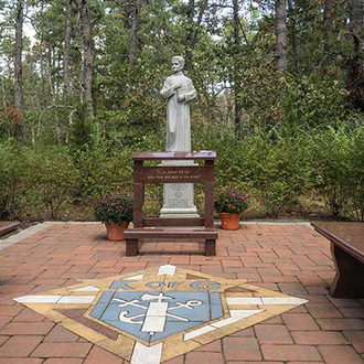 A reflection garden with custom benches and a kneeler adorn a statue of Blessed Michael McGivney on a patio surrounded by trees