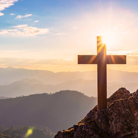 A cross on top of a mountain peak with sunlight shining on it from the background.