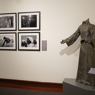 Color photo of a museum gallery with black and white photographs and a decapitated and bullet riddled statue of Christ with his arms outstretched. 