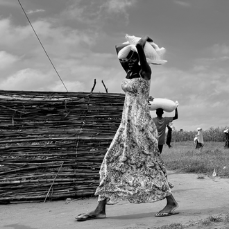 Black and white photo of a woman walking outdoors with a bag of food on her head.