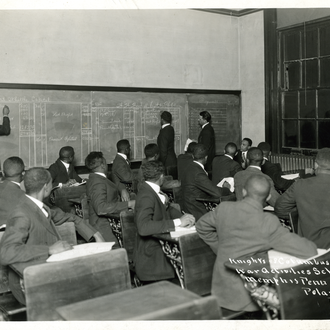 War Activities School bookkeeping class. Men sitting in classroom turn to see exercise on blackboards behind them. Three men stand at the blackboards.