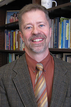 Headshot of Moral Theologian Dr. John Grabowski, who smiles while wearing a tan plaid suit, and a striped tie