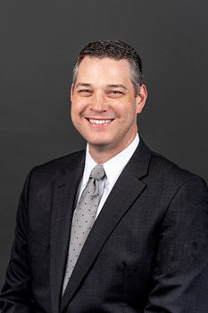 Headshot of James Gaul, who smiles while wearing a black suit and a gray polka dotted tie