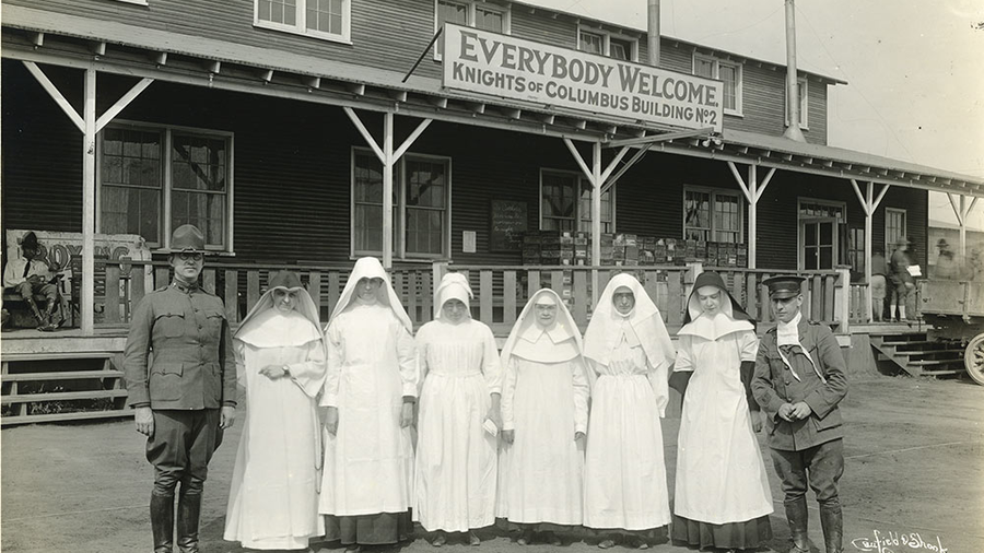 Knights of Columbus secretary Thomas Clines (left) and military chaplain Father Regis Barrett (right) stand with religious sisters who volunteered as nurses at Camp Zachary Taylor, Ky. During the 1918 influenza epidemic, the K of C hut was used as an emergency hospital.