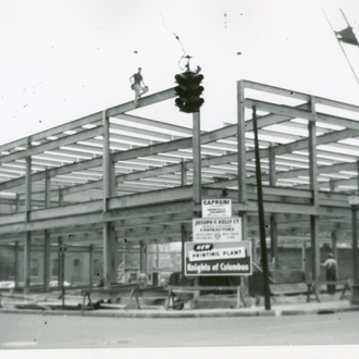 Black and white photo showing Metal framing of a large building, highlighting the name of the building with a large sign out front, "Knights of Columbus Printing Plant."