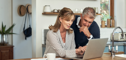A woman sitting at a table pointing to her laptop screen and showing a man, leaning on the table, something on the laptop.