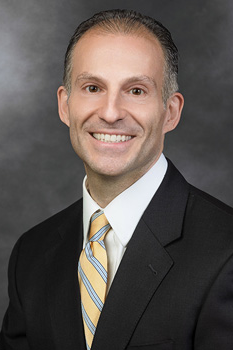 Michael Prinzivalli, who smiles while wearing a black suit and a yellow and blue striped tie