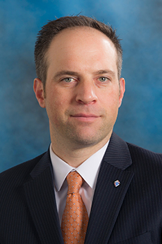 Headshot of Michael Votto, who smiles while wearing a black suit and a burnt orange tie