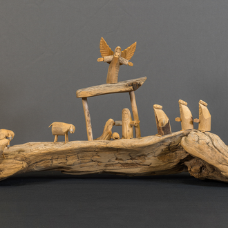 Photo of a crèche featuring the wood carved figures of the Holy Family in the stable, an Angel, the 3 Magi, and 7 sheep on a piece of driftwood. 