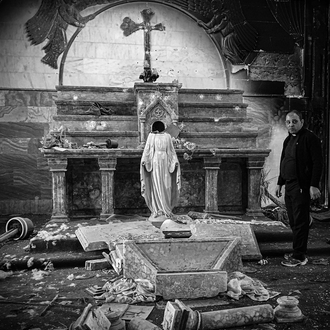 Black and white photo of a priest standing near of the altar inside a damaged church. At center stands a statue of the Blessed Virgin Mary with her face and hands missing.