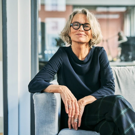 A woman sitting and smiling with gray hair and round glasses.